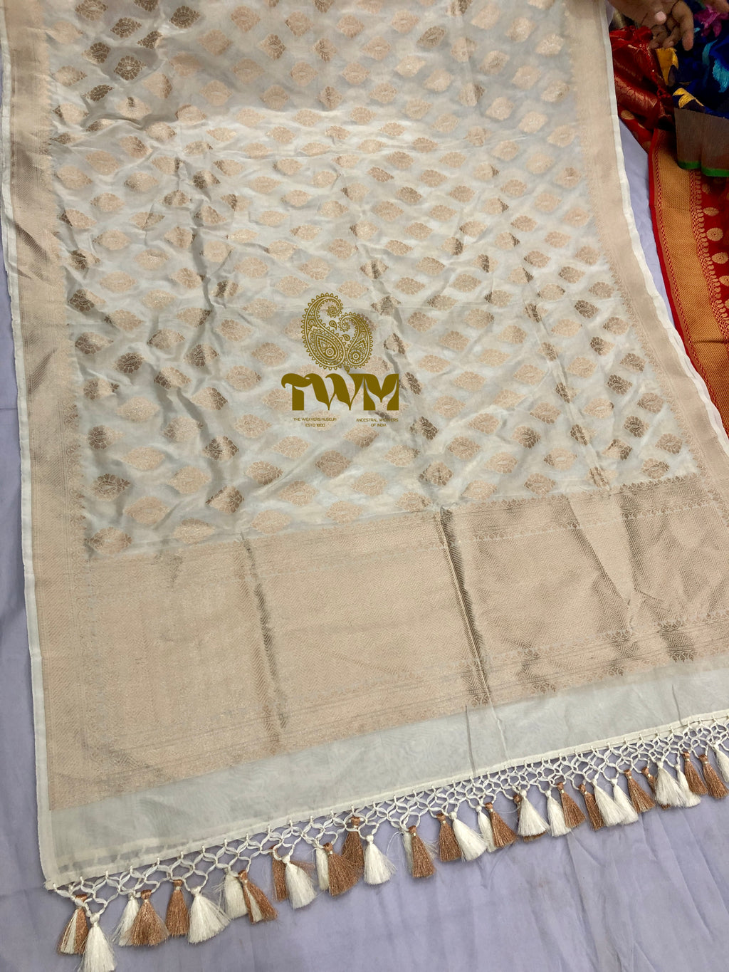Beige fabric with gold patterns and tassels, featuring a logo, on a light gray background.