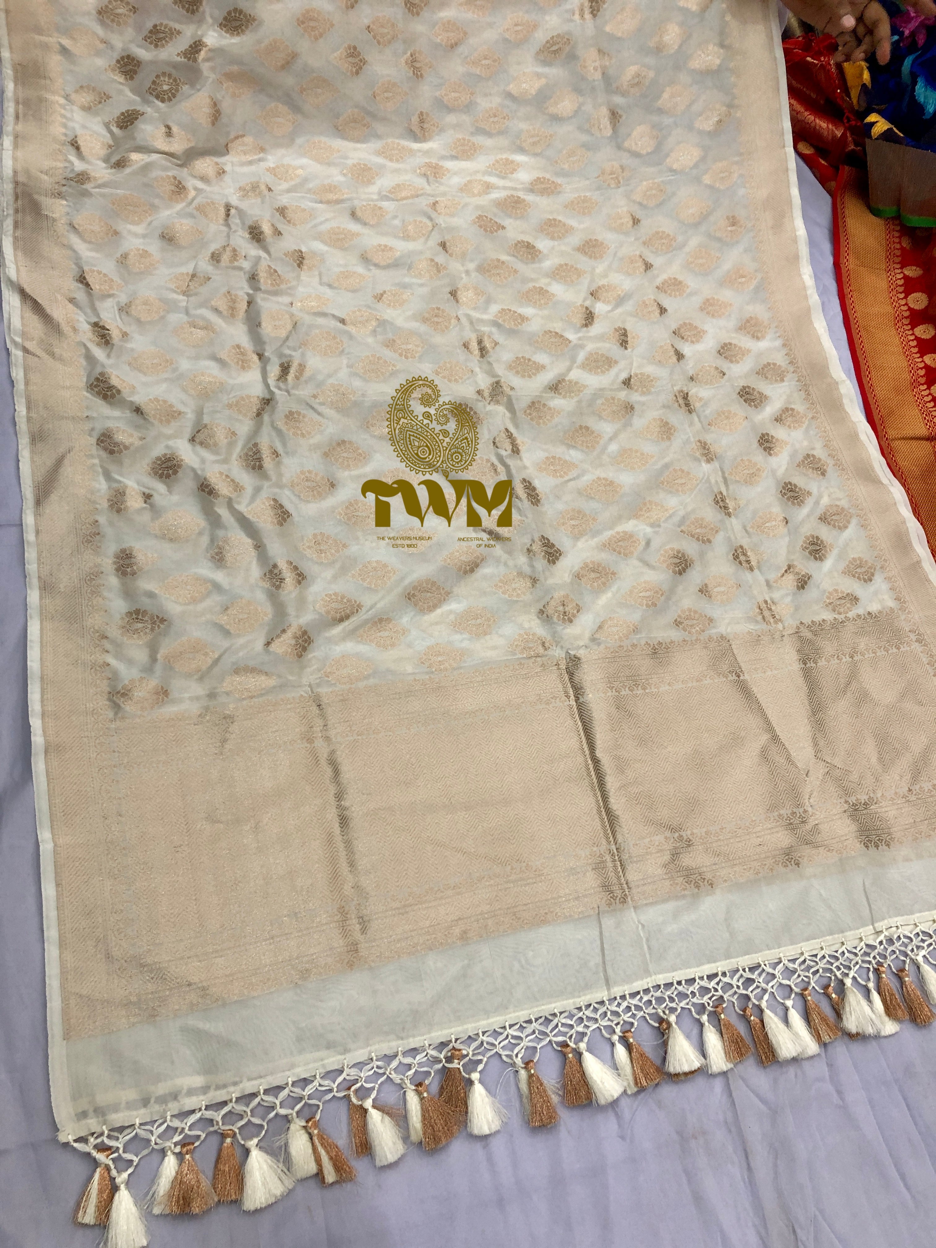 Beige fabric with gold patterns and tassels, featuring a logo, on a light gray background.